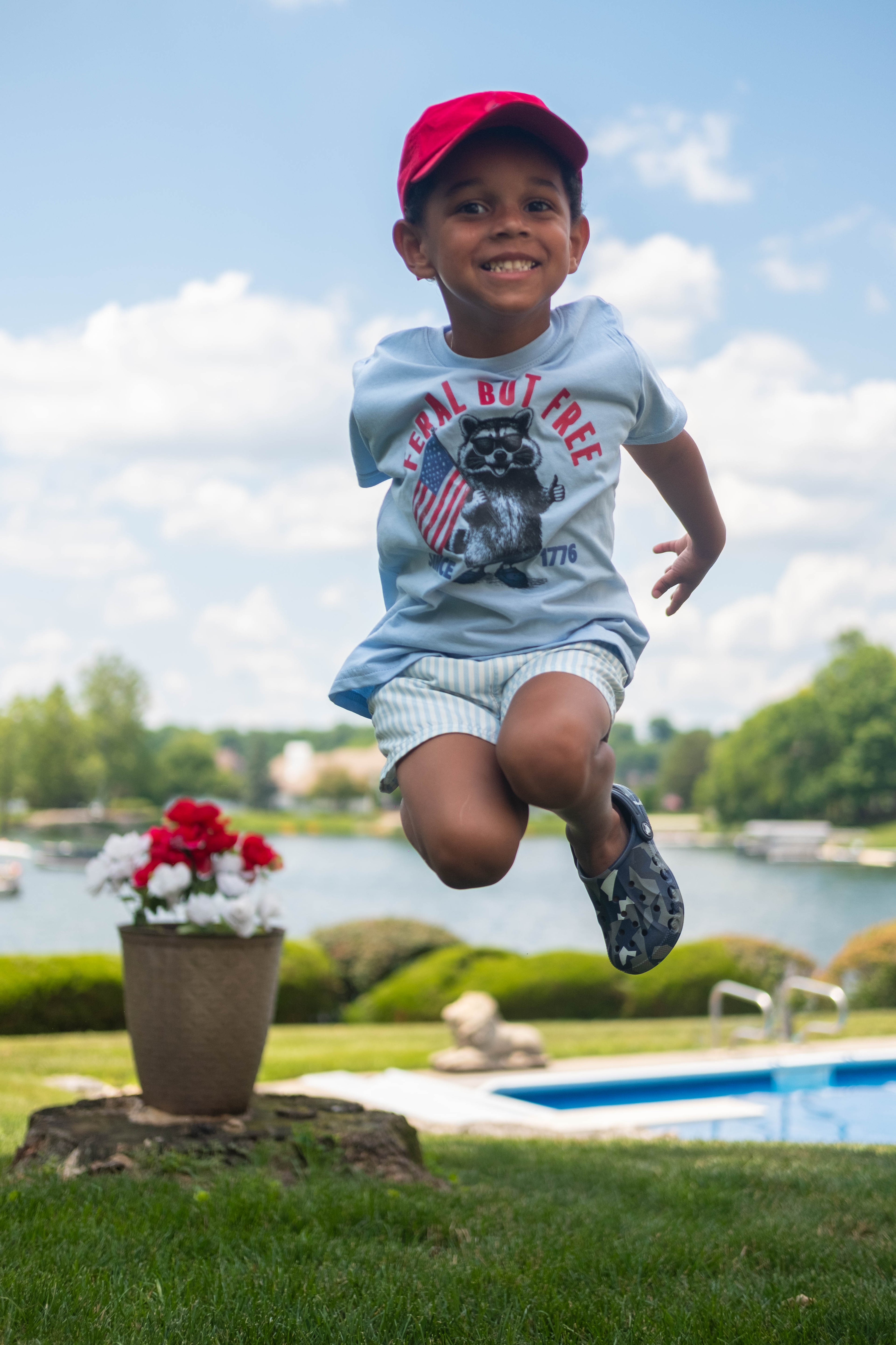 A young boy jumps joyfully outdoors over a swimming pool, wearing a light blue graphic t-shirt, white shorts, a red baseball cap, and dark sneakers. The background shows a sunny day with a lake, trees, and a blue sky.